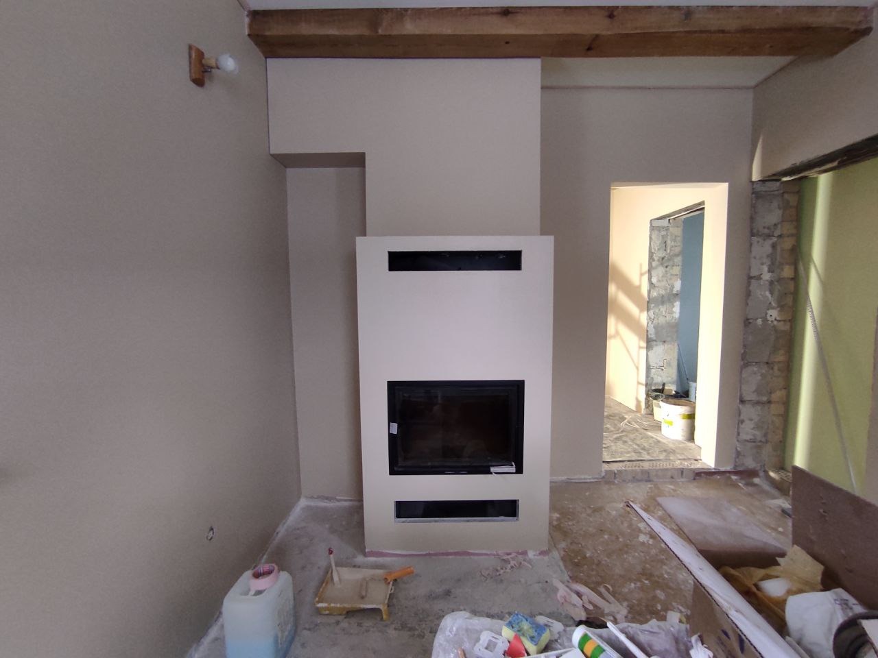 Wnętrze w trakcie prac wykończeniowych, widoczny obudowany kominek z czarnym wkładem, ściany przygotowane do malowania, surowy drewniany belkowy sufit, podłoga zabezpieczona folią, widoczne...