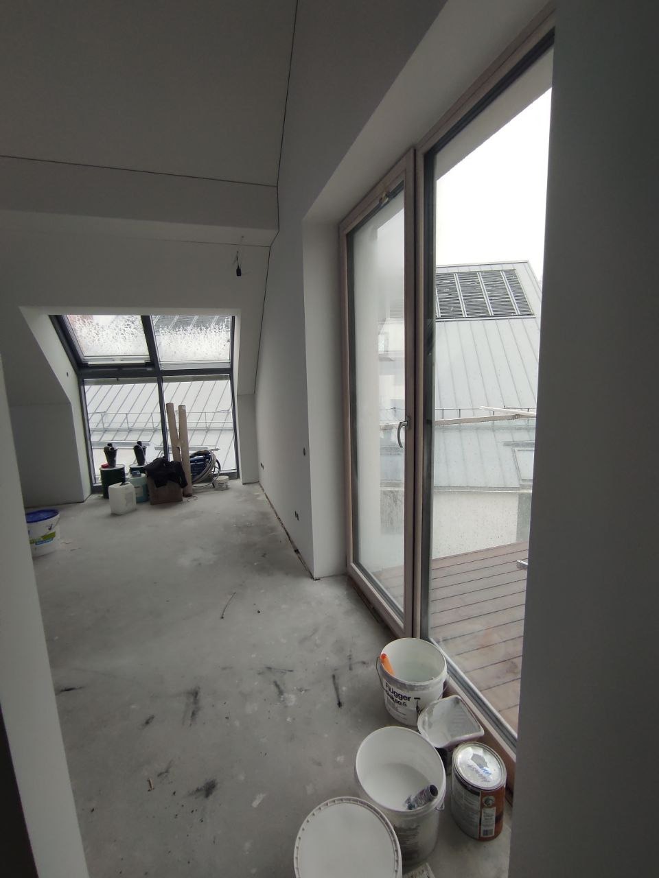 Wnętrze w trakcie prac przygotowawczych: surowe ściany, okna dachowe, widoczne wiadra z farbą i narzędzia, podłoga zabezpieczona przed zabrudzeniem.