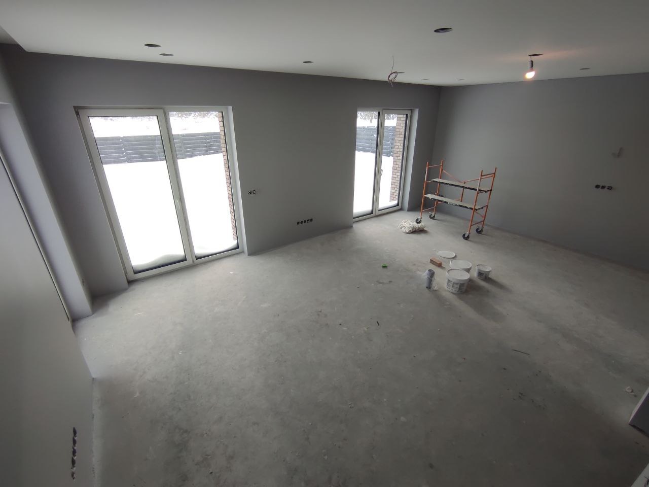 Wnętrze pomalowanego na szaro pokoju w trakcie wykańczania; widoczne okna z widokiem na ośnieżony ogród, metalowy regał na kółkach, puszki z farbą i niedoskonałości na podłodze.