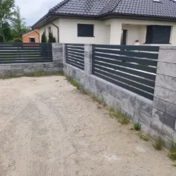 Nowoczesne ogrodzenie z szarych paneli poziomych i betonowych słupków, widok z zewnątrz posesji, na tle domu z ciemną dachówką.