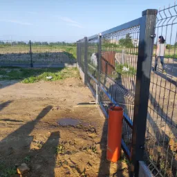 Fragment nowo zamontowanego, czarnego ogrodzenia panelowego z widoczną bramą wjazdową, na nierównym, piaszczystym terenie z kałużą; w tle pole i częściowo widoczna sylwetka kobiety.