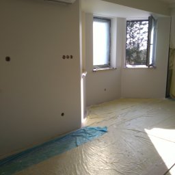 AUTOFIX - Pokój w trakcie przygotowań do prac wykończeniowych, podłoga i okna zabezpieczone folią ochronną, widoczne puszki instalacyjne na ścianie, klimatyzator zakryty folią.