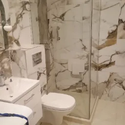 Wykończona łazienka z kabiną typu walk-in, marmurowymi płytkami na ścianach, białą armaturą i chromowaną deszczownicą. Nowoczesne i eleganckie wnętrze.