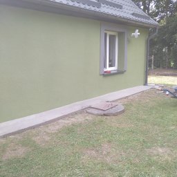 AUTOFIX - Budynek z ocieploną i pomalowaną elewacją na zielono. Widoczne okno z szarą ramą, szary dach i fragment trawnika. Na parapecie leżą niewielkie przedmioty.