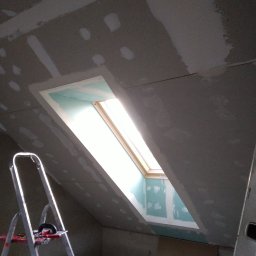 AUTOFIX - Poddasze z oknem dachowym w trakcie montażu płyt gipsowo-kartonowych, widoczna drabina i narzędzia. Szpachlowanie łączeń płyt na skośnej ścianie.