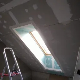 Poddasze z oknem dachowym w trakcie montażu płyt gipsowo-kartonowych, widoczna drabina i narzędzia. Szpachlowanie łączeń płyt na skośnej ścianie.