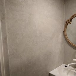 AUTOFIX - Ściana w łazience z dekoracyjną, szarą strukturą tynku w stylu betonu, z fragmentem lustra w ramie z liny jutowej i umywalką w rogu.