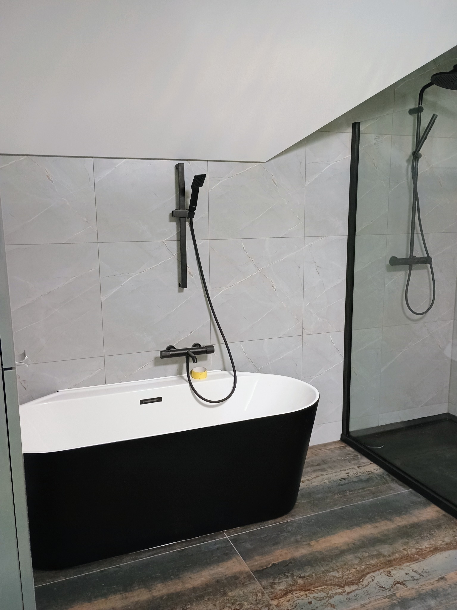 Nowoczesna łazienka z wolnostojącą wanną w kolorze czarno-białym, czarną armaturą i kabiną prysznicową z czarnymi ramami, wykończona szarymi płytkami imitującymi marmur i podłogą w odcieniach...