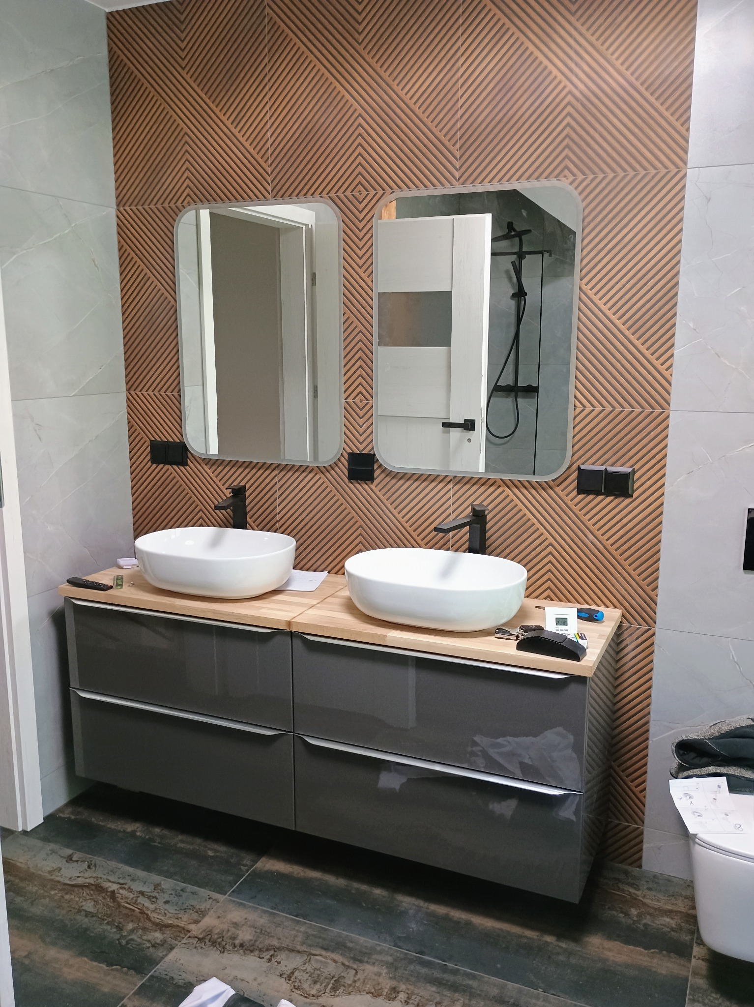 Nowoczesna łazienka z dwoma owalnymi umywalkami nablatowymi, szarą szafką z szufladami, drewnianym blatem, geometryczną okładziną ścienną w odcieniach brązu i szaro-białymi płytkami imitującymi...