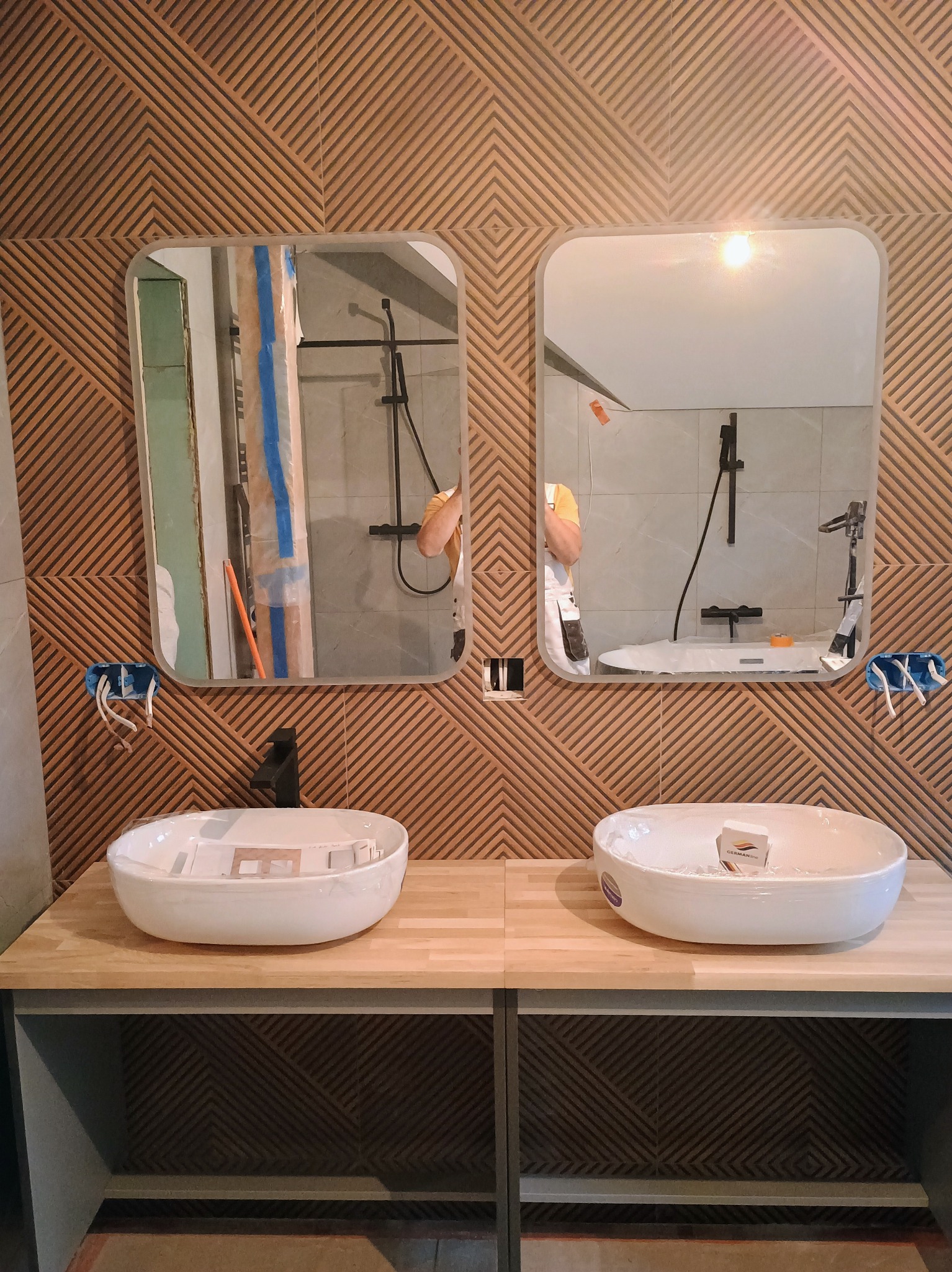 Dwie białe, owalne umywalki nablatowe na drewnianym blacie w łazience z geometryczną ścianą. W lustrach odbicie kabiny prysznicowej z czarną armaturą. Widoczne wystające kable elektryczne.