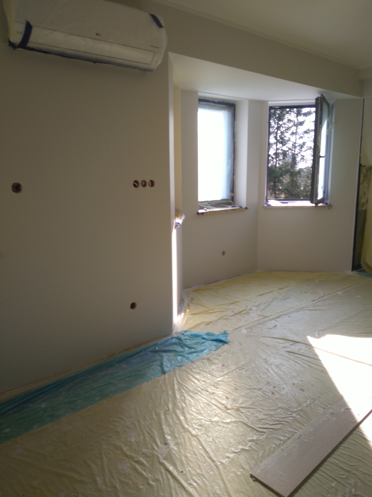 Pokój w trakcie przygotowań do prac wykończeniowych, podłoga i okna zabezpieczone folią ochronną, widoczne puszki instalacyjne na ścianie, klimatyzator zakryty folią.