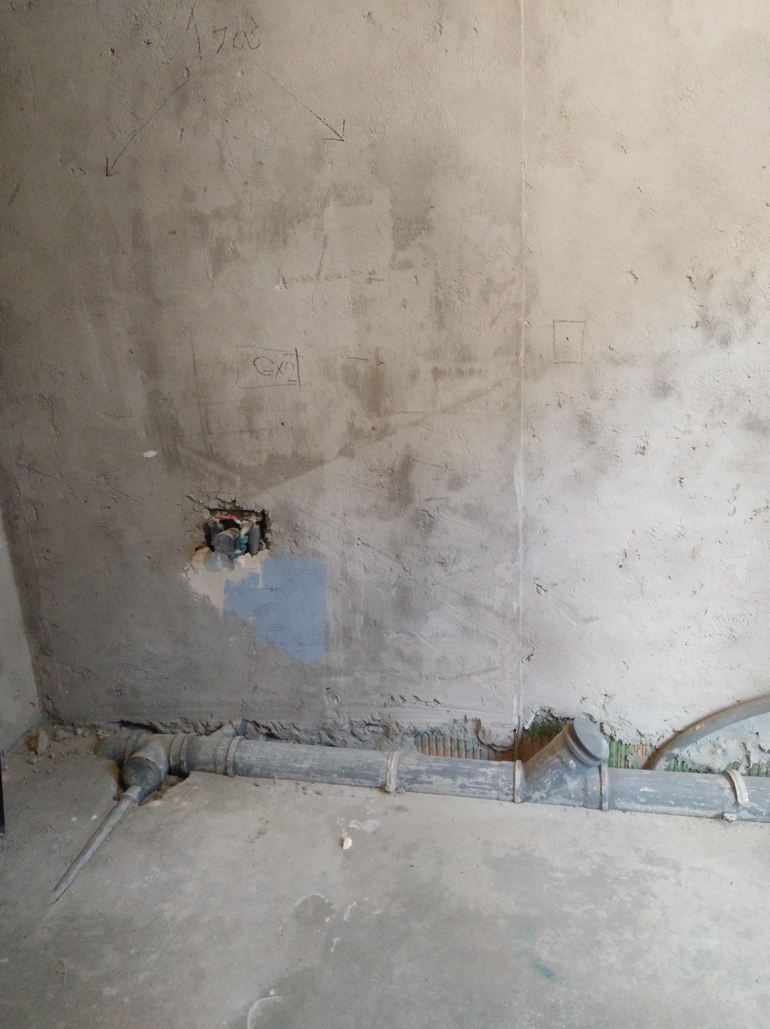 Surowa ściana w trakcie prac instalacyjnych w łazience z widocznymi rurami kanalizacyjnymi, otworem na instalację elektryczną i odręcznymi notatkami na ścianie.