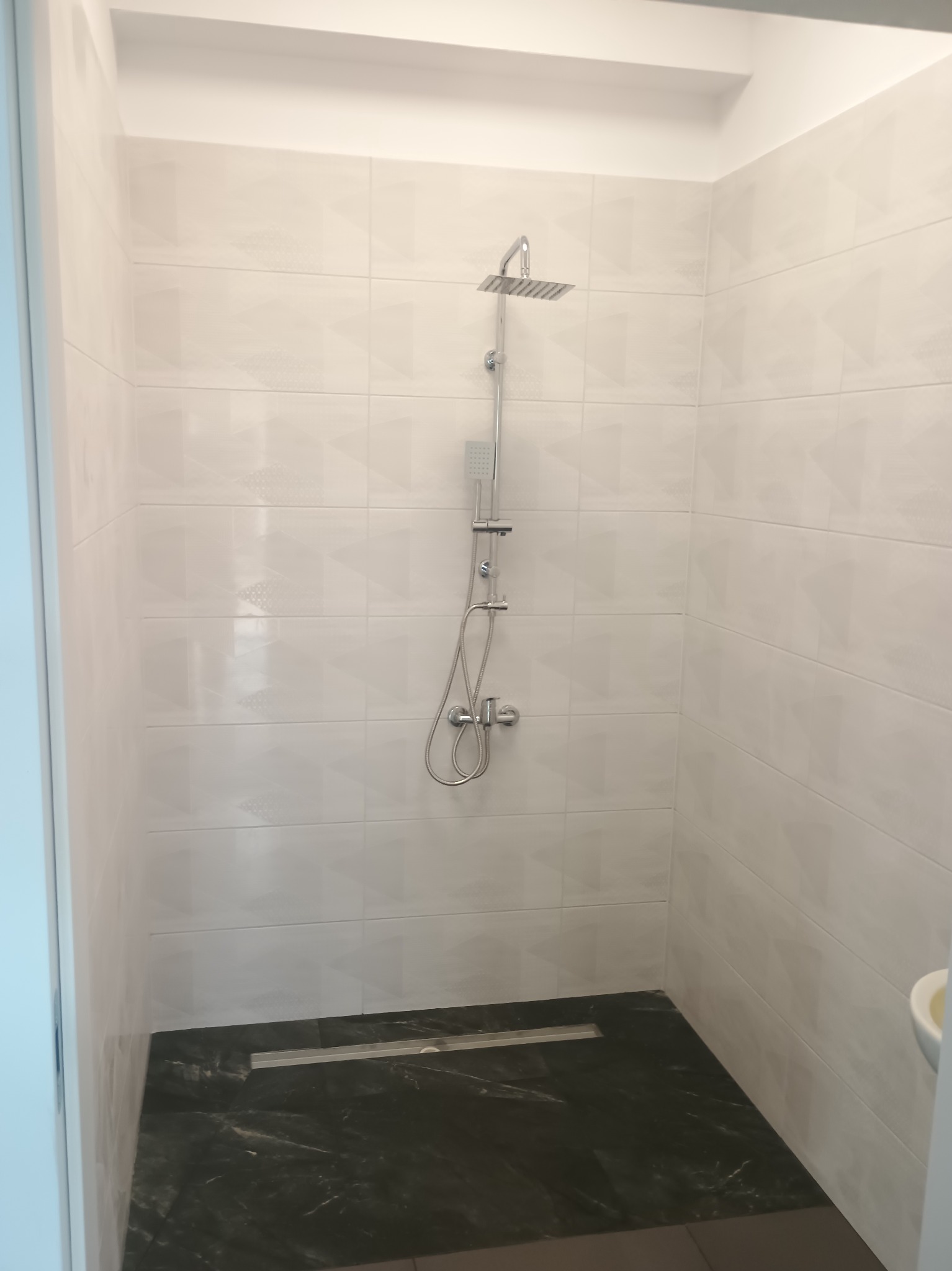 Nowoczesna kabina prysznicowa z odpływem liniowym, wykończona białymi płytkami z subtelnym wzorem i ciemną, marmurkową posadzką. Minimalistyczny design.