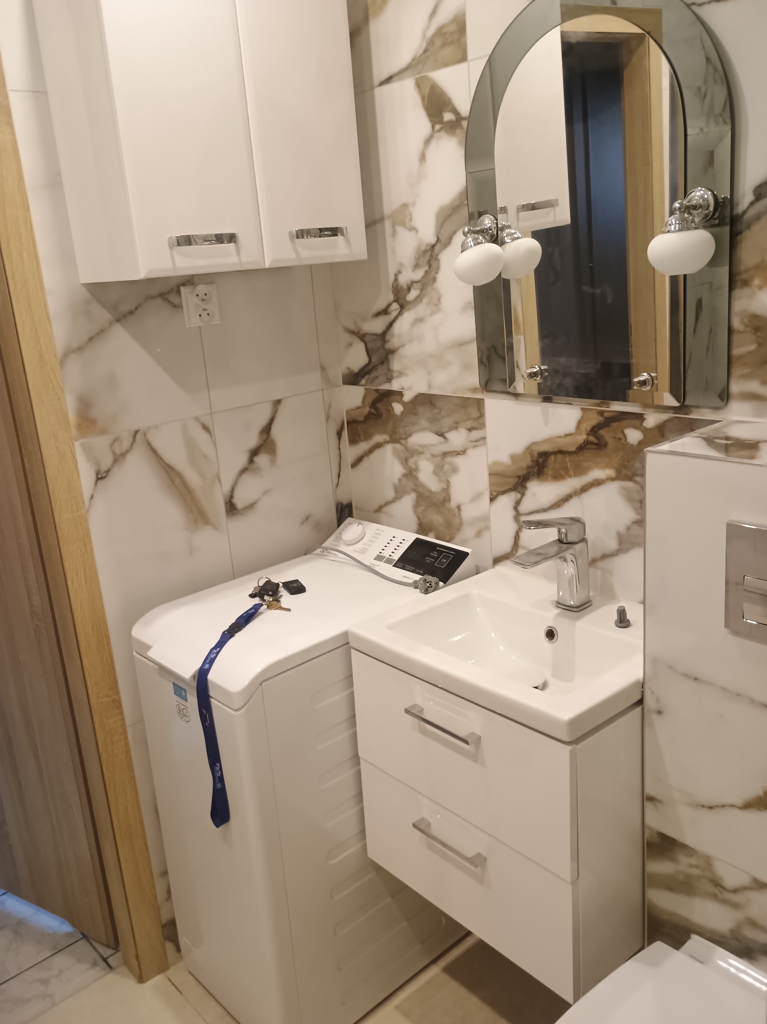 Nowoczesna łazienka z marmurowymi płytkami, umywalką z szafką i pralką. Lustro owalne z oświetleniem. Klucze leżą na pralce.