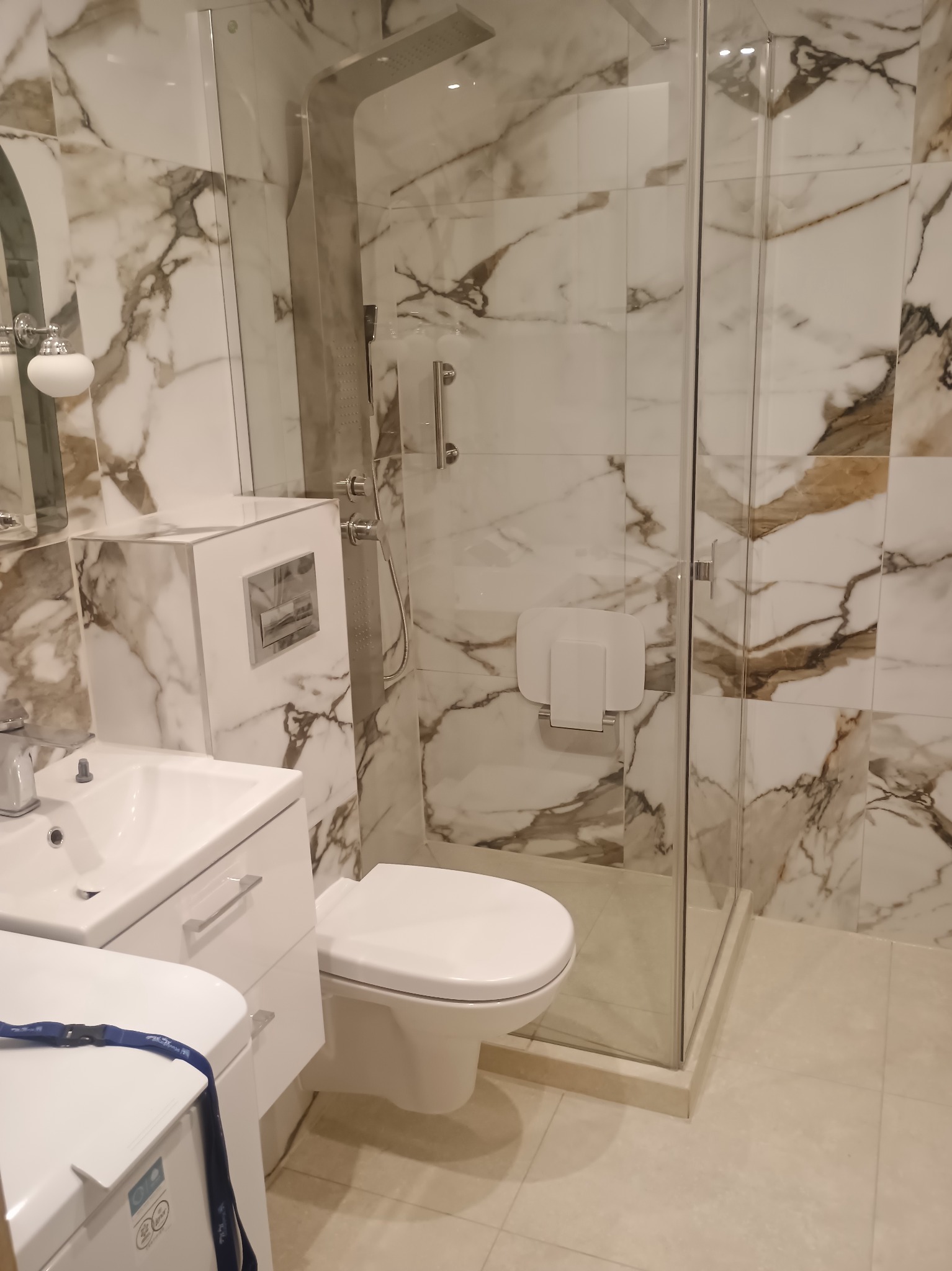 Wykończona łazienka z kabiną typu walk-in, marmurowymi płytkami na ścianach, białą armaturą i chromowaną deszczownicą. Nowoczesne i eleganckie wnętrze.