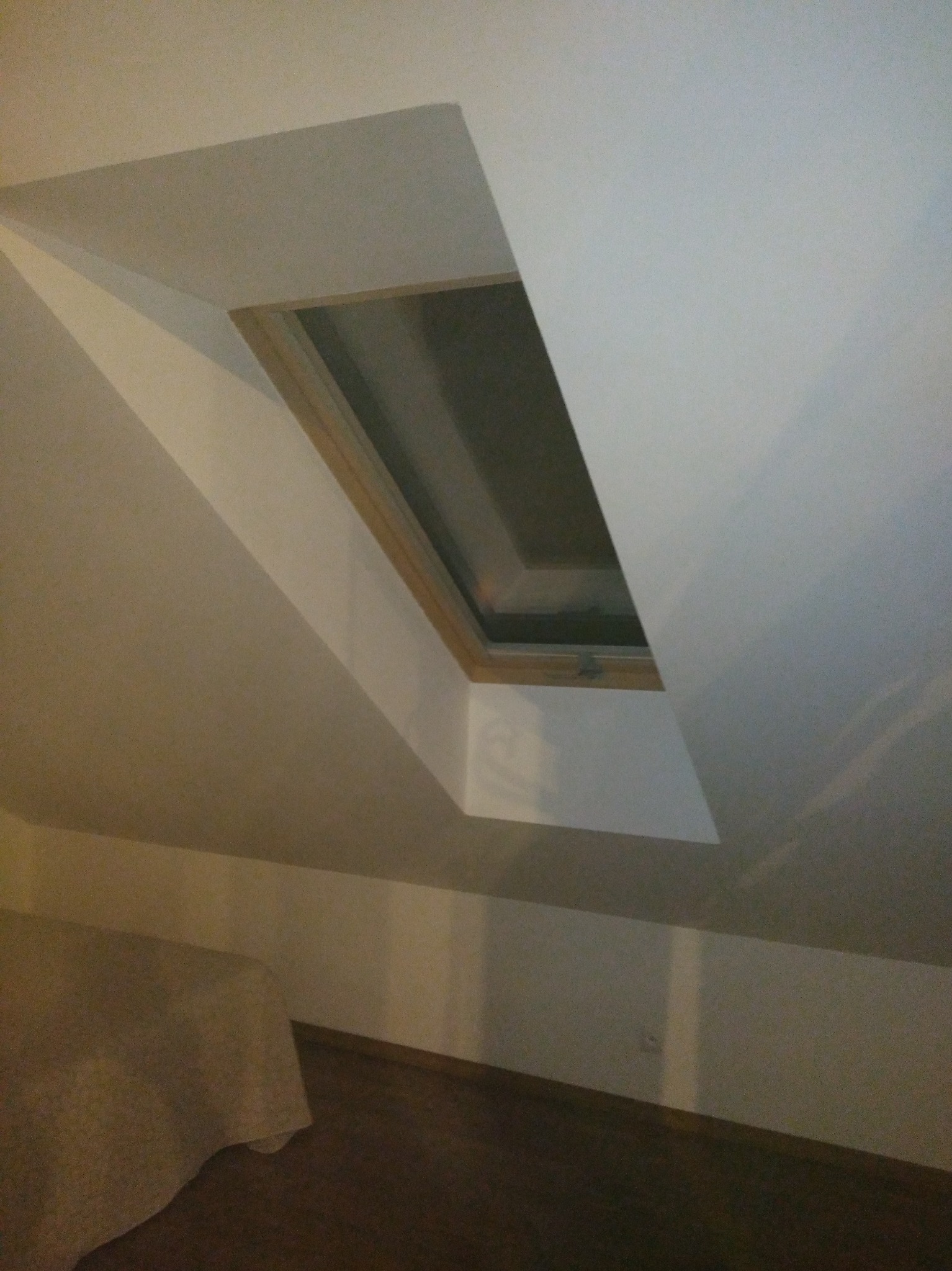 Poddasze z oknem dachowym w drewnianej ramie, świeżo po malowaniu na biało. Widoczny fragment podłogi i łóżka. Ujęcie z dołu.