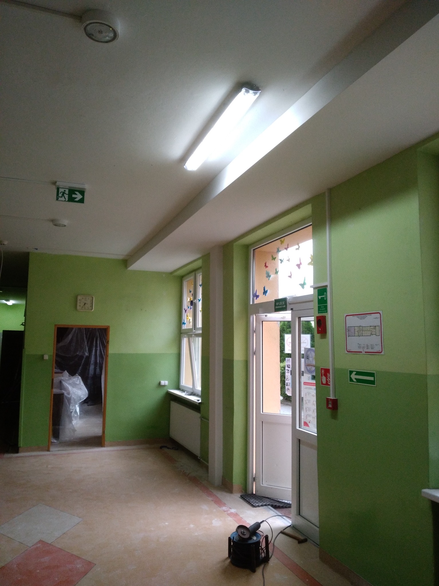 Wnętrze w trakcie odświeżania: zielone ściany, widoczne okna z dekoracjami, podłoga z fragmentami płytek i narzędzie do cięcia. Oświetlenie sufitowe.