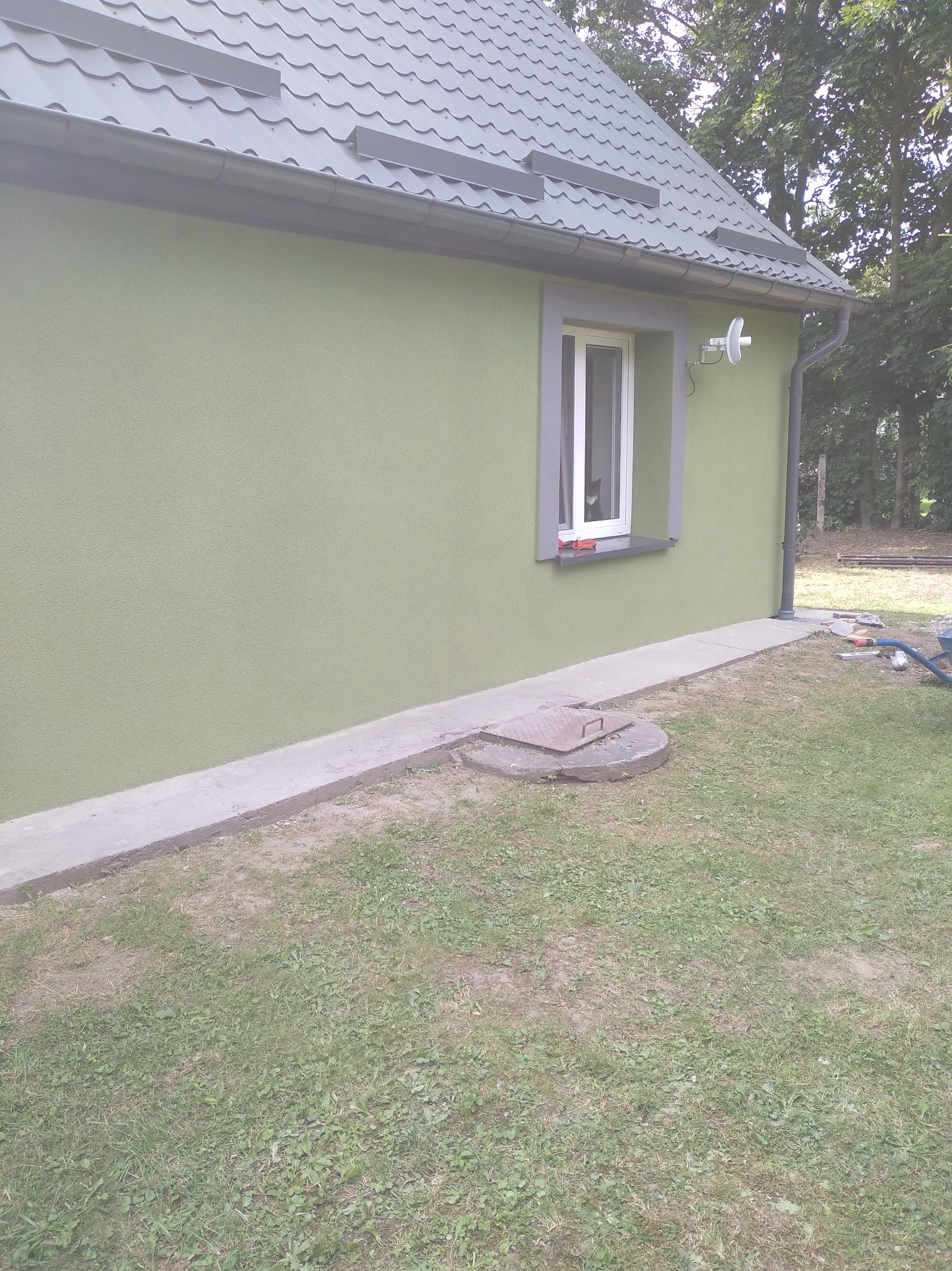Budynek z ocieploną i pomalowaną elewacją na zielono. Widoczne okno z szarą ramą, szary dach i fragment trawnika. Na parapecie leżą niewielkie przedmioty.