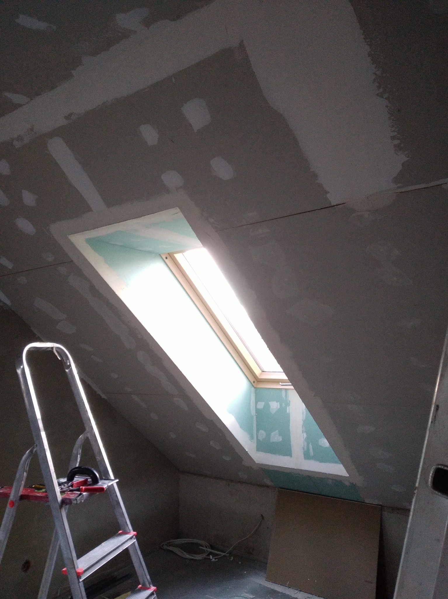 Poddasze z oknem dachowym w trakcie montażu płyt gipsowo-kartonowych, widoczna drabina i narzędzia. Szpachlowanie łączeń płyt na skośnej ścianie.