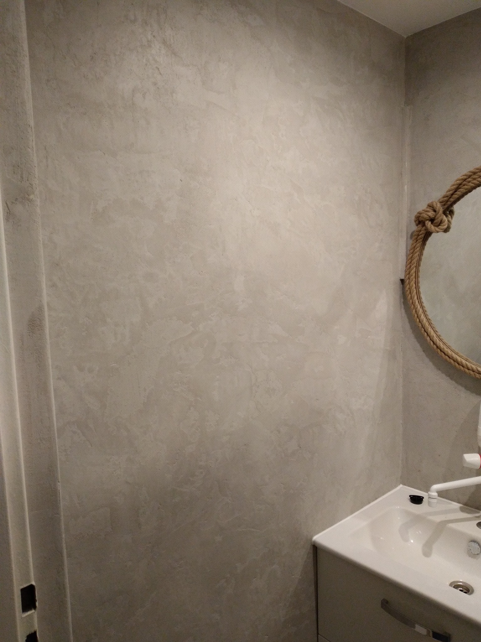 Ściana w łazience z dekoracyjną, szarą strukturą tynku w stylu betonu, z fragmentem lustra w ramie z liny jutowej i umywalką w rogu.