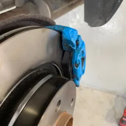 Szczegółowy widok tarczy hamulcowej z niebieskim zaciskiem i nowymi klockami hamulcowymi, widoczne ślady korozji na piaście koła.