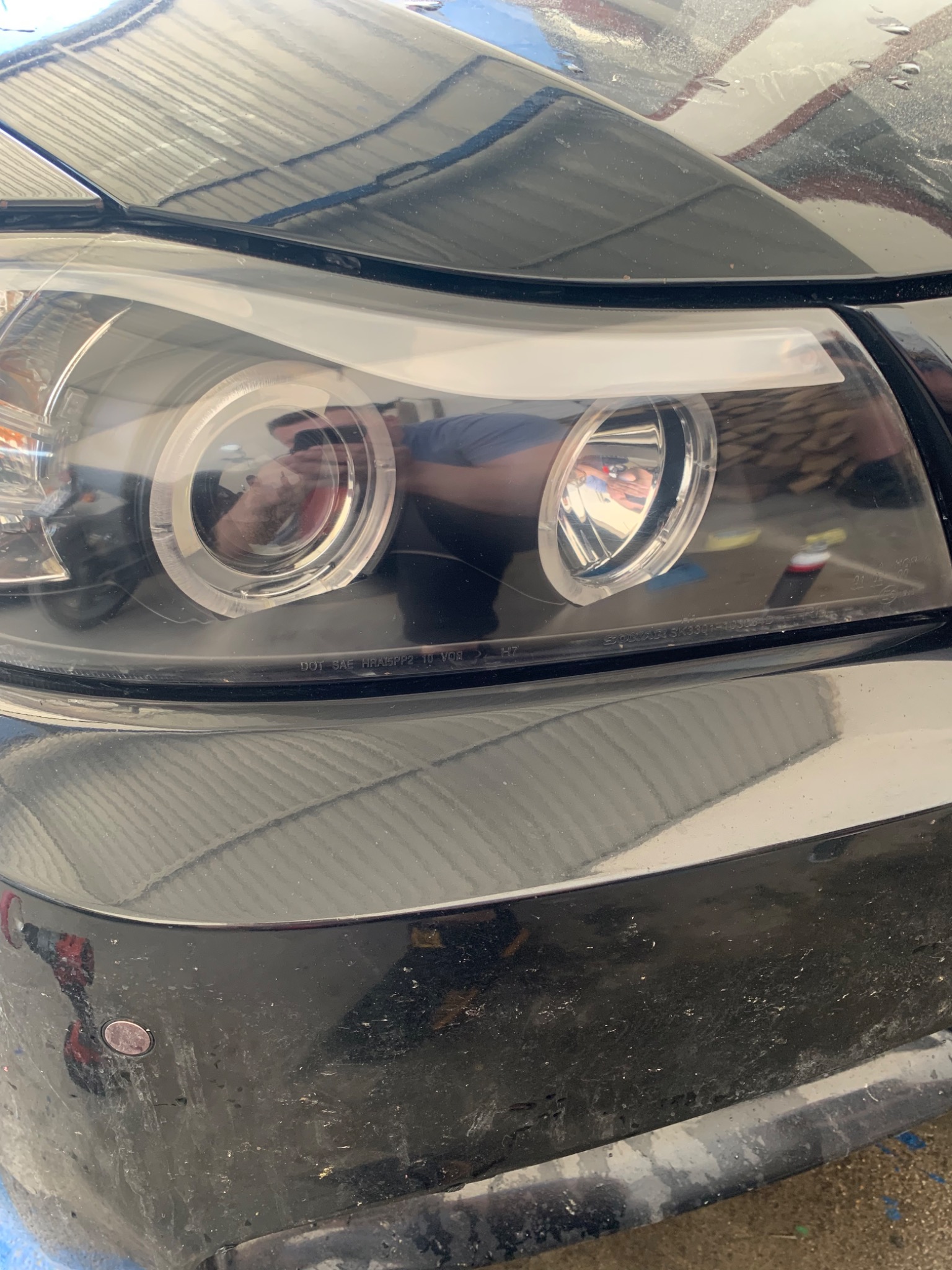 Zbliżenie na przedni reflektor samochodu z widocznymi śladami zabrudzeń na karoserii i odbiciem osoby robiącej zdjęcie w soczewce lampy.