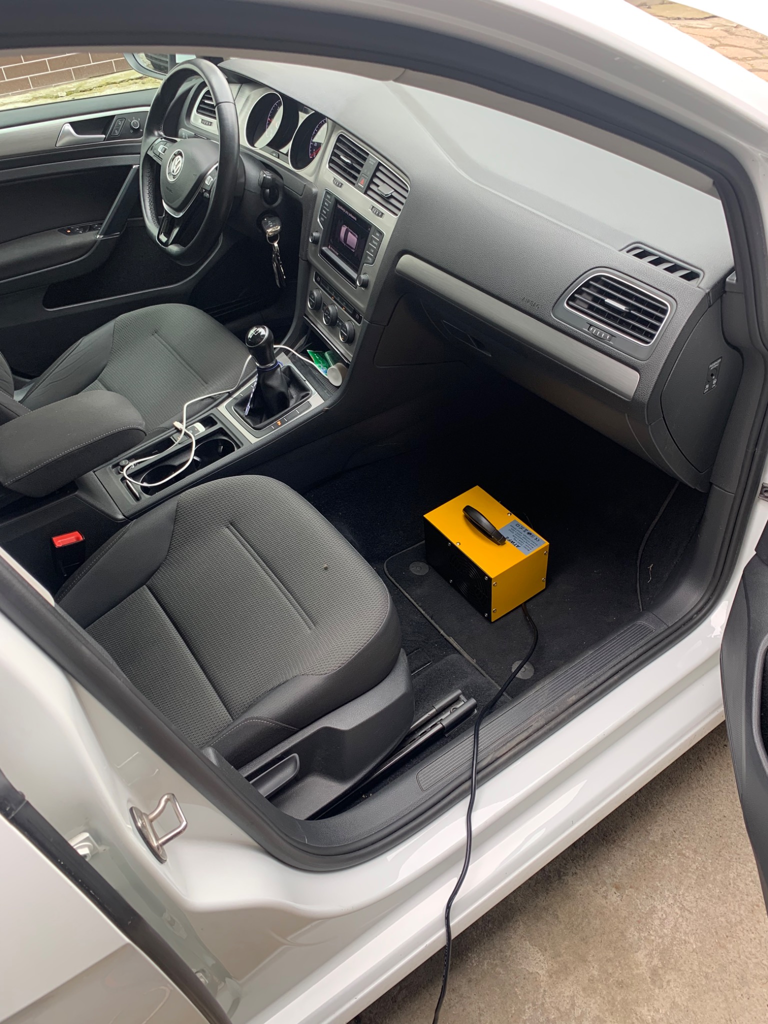 Wnętrze samochodu Volkswagen Golf z otwartymi drzwiami, widoczny żółty generator ozonu na dywaniku pasażera, prawdopodobnie w trakcie odgrzybiania klimatyzacji.