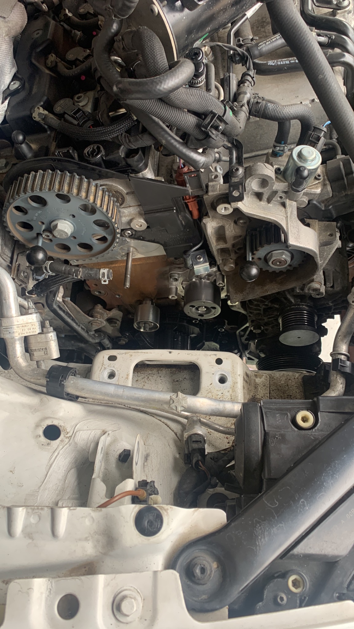 Widok z góry na silnik samochodowy w trakcie naprawy, z widocznymi elementami rozrządu, przewodami i osprzętem. Zauważalny brud i ślady eksploatacji na elementach.