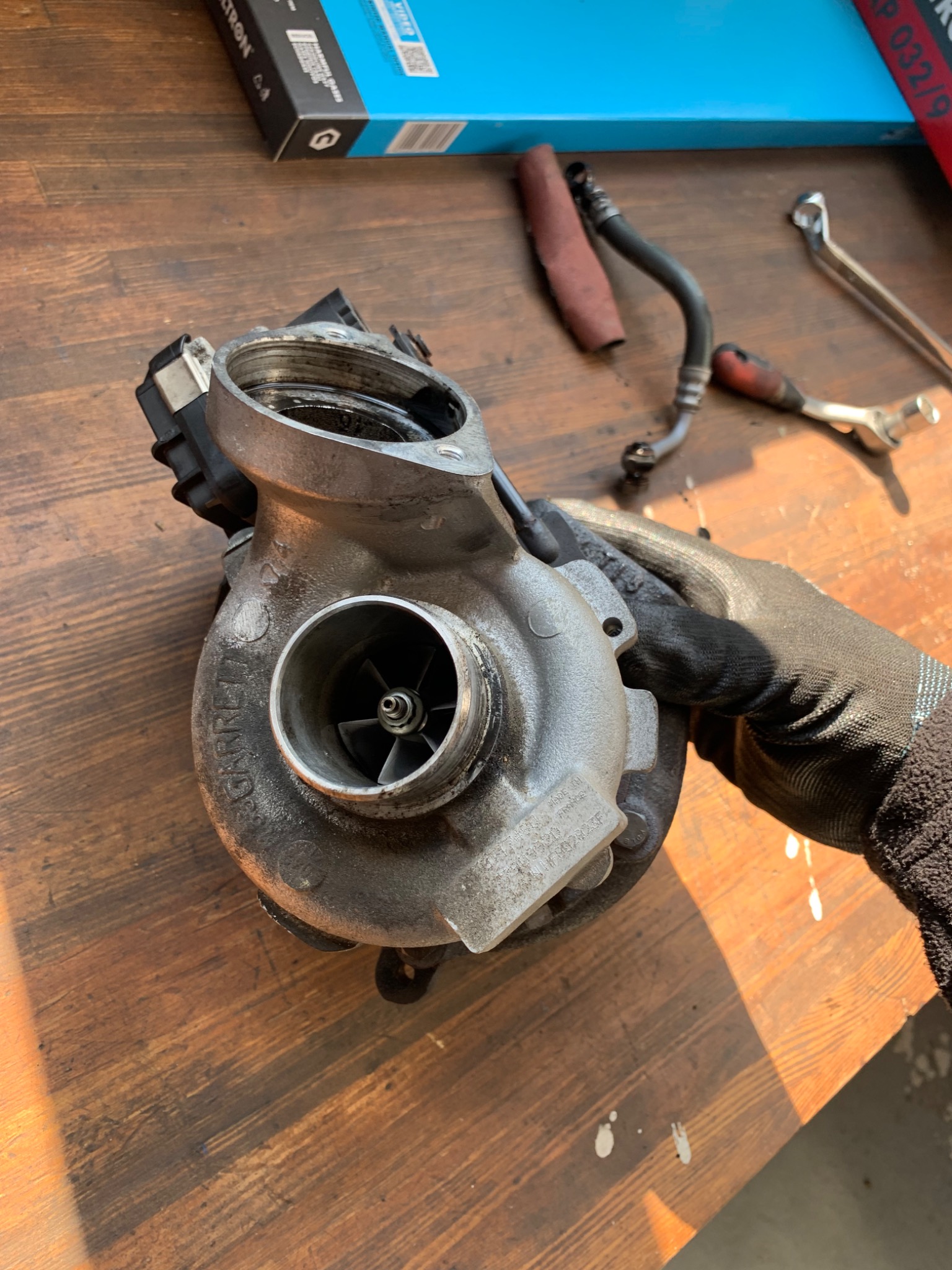 Turbosprężarka z widocznymi śladami użytkowania, trzymana w rękawicy roboczej na drewnianym stole warsztatowym, obok leżą klucze i przewody.