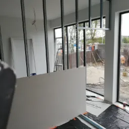 Wnętrze w trakcie montażu ścianki działowej z płyt kartonowo-gipsowych na metalowym stelażu, widoczne profile, płyty, folia ochronna na podłodze i okna z widokiem na plac budowy.