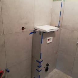 Urban - Ściany w łazience wyłożone szarymi płytkami, widoczny stelaż podtynkowy toalety obłożony płytami, niebieska taśma zabezpieczająca narożniki, wystające przewody elektryczne.