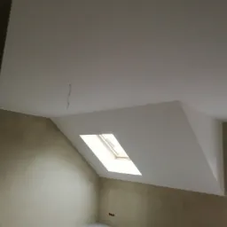 Poddasze po szpachlowaniu z oknem dachowym, widoczne gładkie ściany przygotowane do malowania, wystający kabel z sufitu i niezabezpieczone rury przy podłodze.