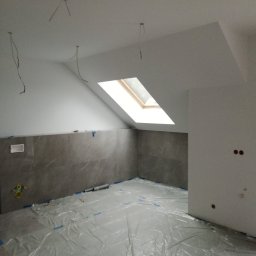 Urban - Poddasze w trakcie prac wykończeniowych: szare płytki imitujące marmur na ścianie, zabezpieczona folią podłoga, wystające kable z sufitu, okno dachowe i przygotowane otwory pod gniazdka elektryczne.
