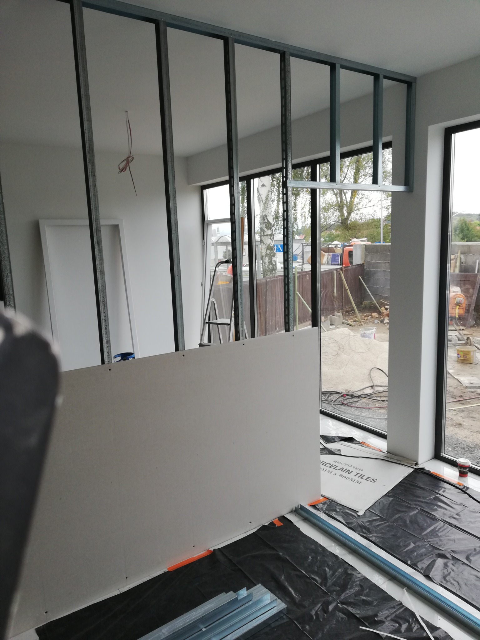 Wnętrze w trakcie montażu ścianki działowej z płyt kartonowo-gipsowych na metalowym stelażu, widoczne profile, płyty, folia ochronna na podłodze i okna z widokiem na plac budowy.