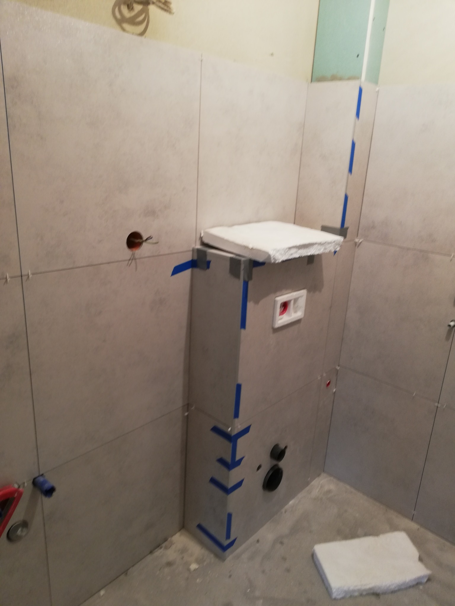 Ściany w łazience wyłożone szarymi płytkami, widoczny stelaż podtynkowy toalety obłożony płytami, niebieska taśma zabezpieczająca narożniki, wystające przewody elektryczne.