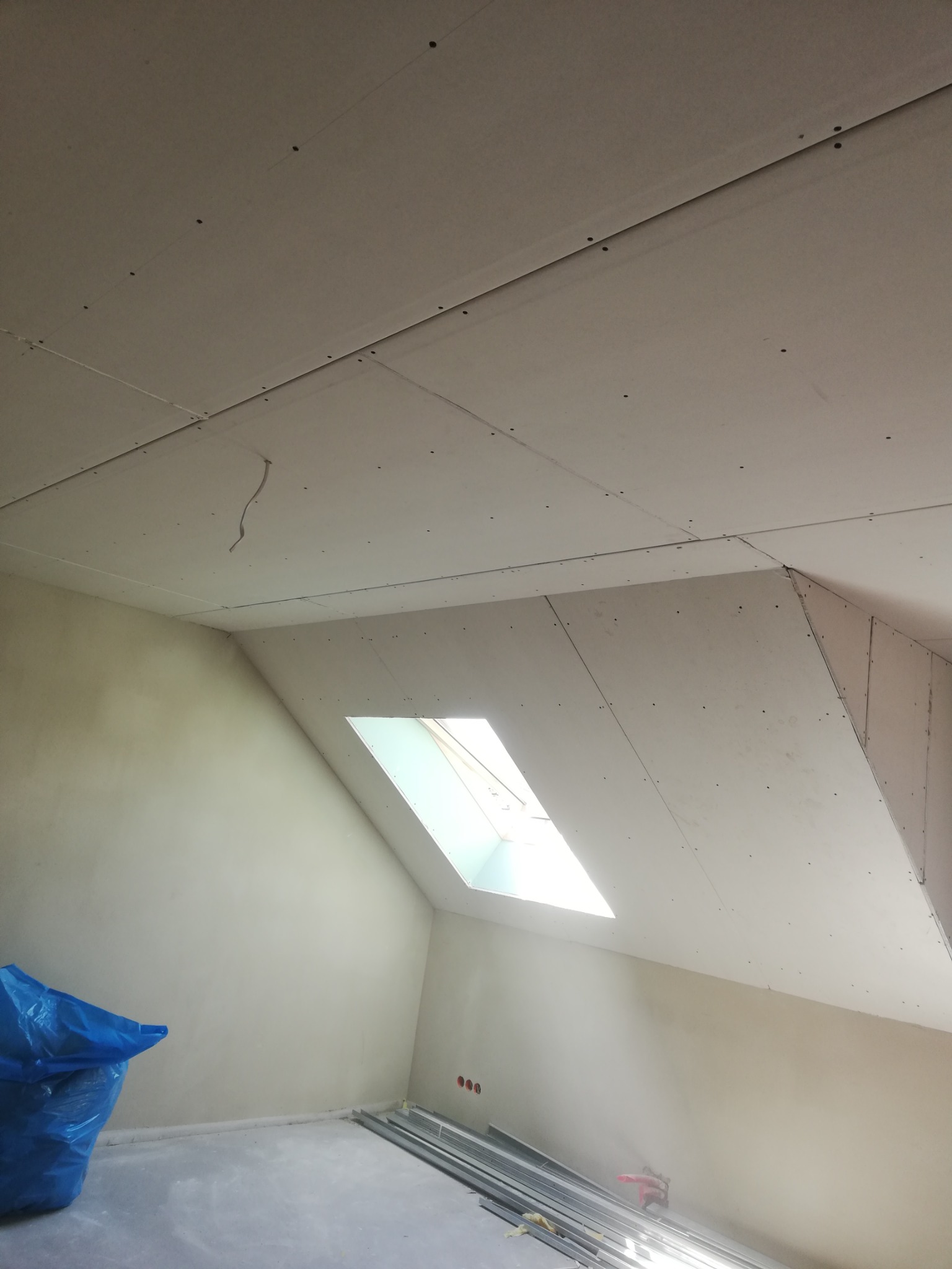 Montaż płyt gipsowo-kartonowych na poddaszu, widoczne łączenia i wkręty, okno dachowe w skosie, profile metalowe na podłodze, niebieski worek z odpadami.