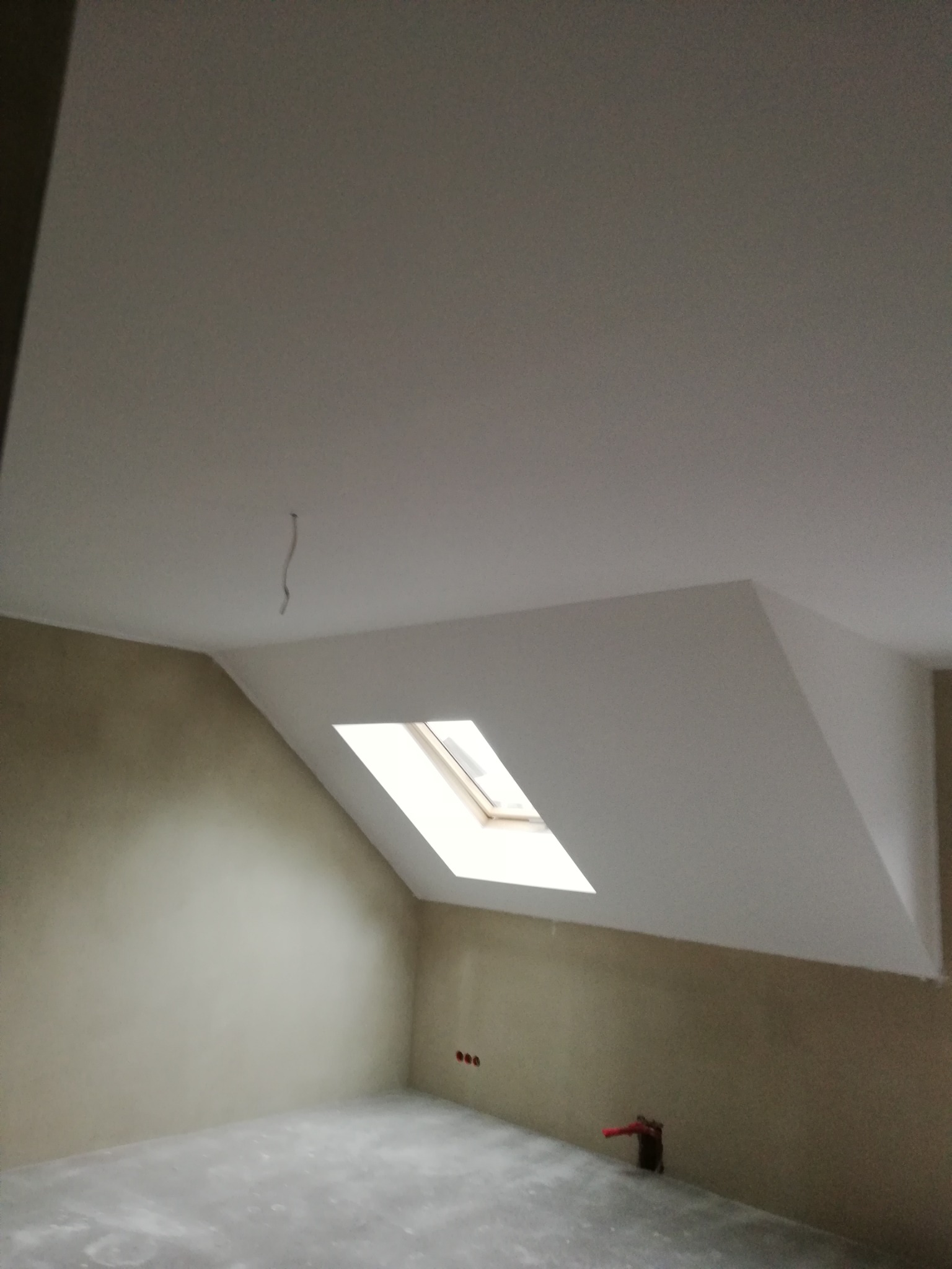 Poddasze po szpachlowaniu z oknem dachowym, widoczne gładkie ściany przygotowane do malowania, wystający kabel z sufitu i niezabezpieczone rury przy podłodze.