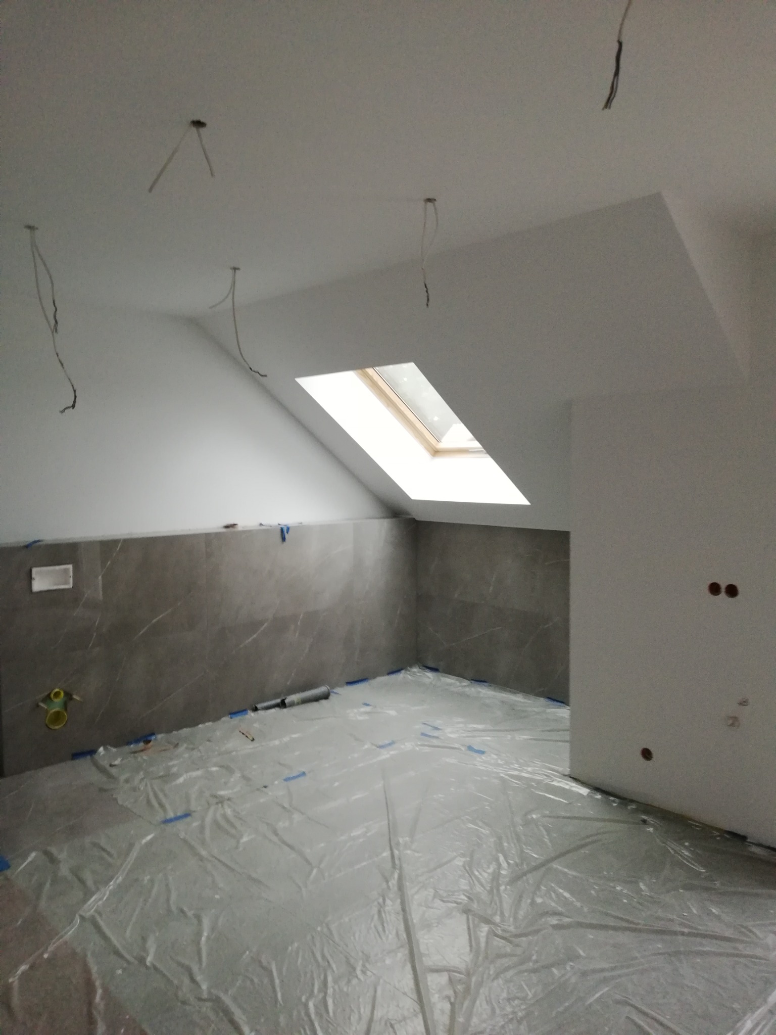 Poddasze w trakcie prac wykończeniowych: szare płytki imitujące marmur na ścianie, zabezpieczona folią podłoga, wystające kable z sufitu, okno dachowe i przygotowane otwory pod gniazdka elektryczne.