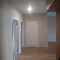 Jasne wnętrze korytarza po odświeżeniu, z białymi drzwiami i ościeżnicami, parkiet ułożony w jodełkę, ściany pomalowane na beżowo, widoczne gniazdko elektryczne.