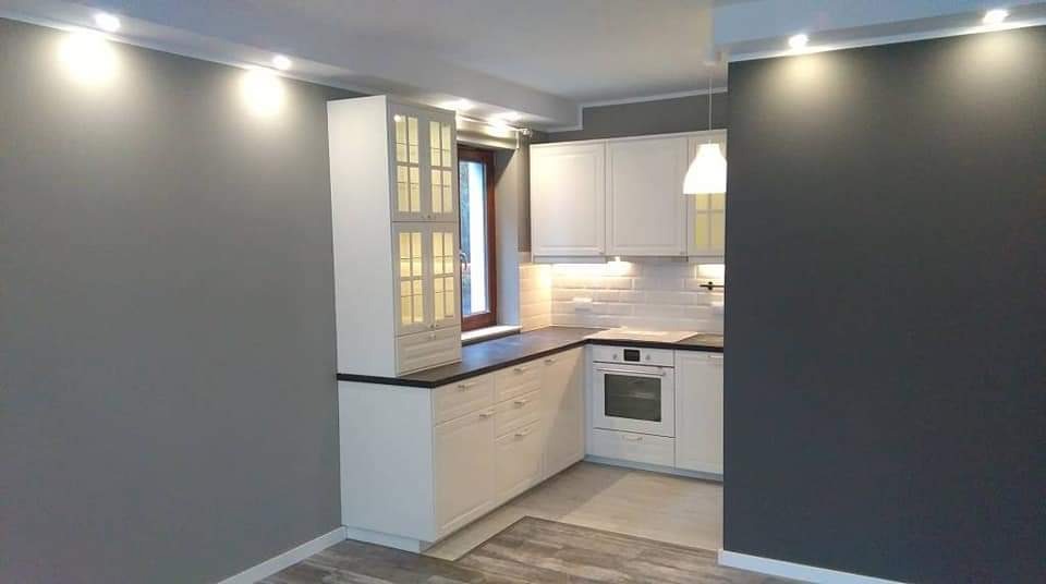 Nowoczesna kuchnia z białymi szafkami z przeszklonymi witrynami, ciemnym blatem, białymi płytkami na ścianie i szarą ścianą w tle, oświetlona punktowymi lampami sufitowymi i wiszącą lampą.