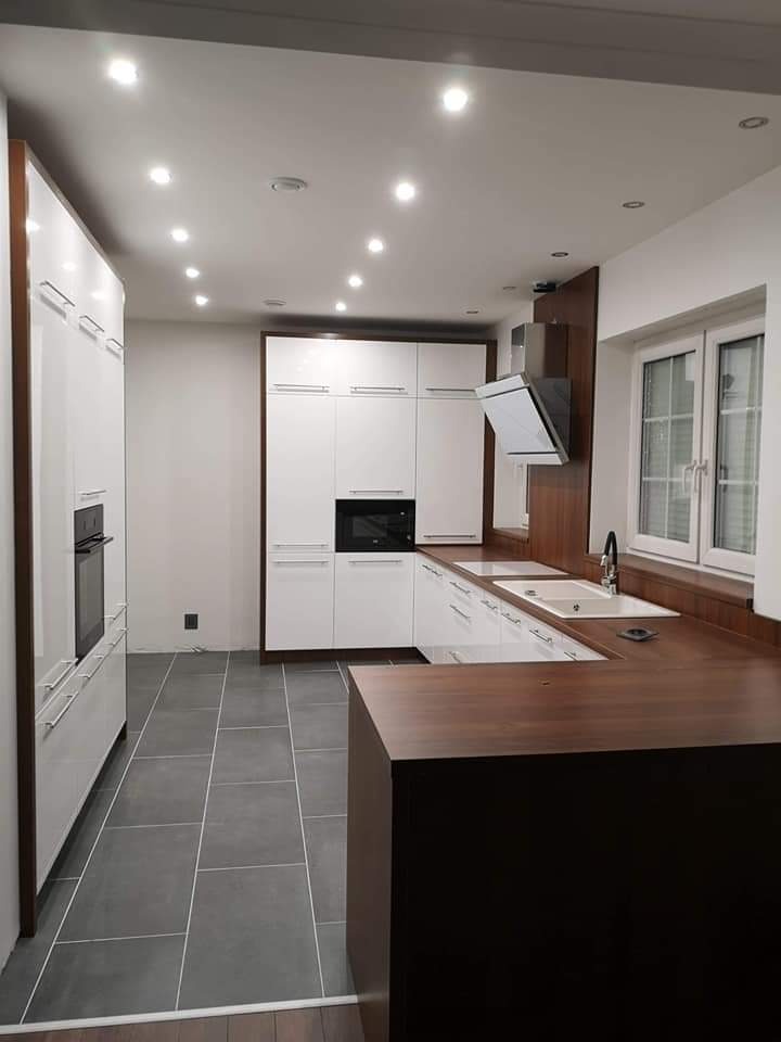 Nowoczesna kuchnia z białymi szafkami, ciemnobrązowym blatem i szarymi płytkami podłogowymi. Oświetlenie punktowe w suficie.