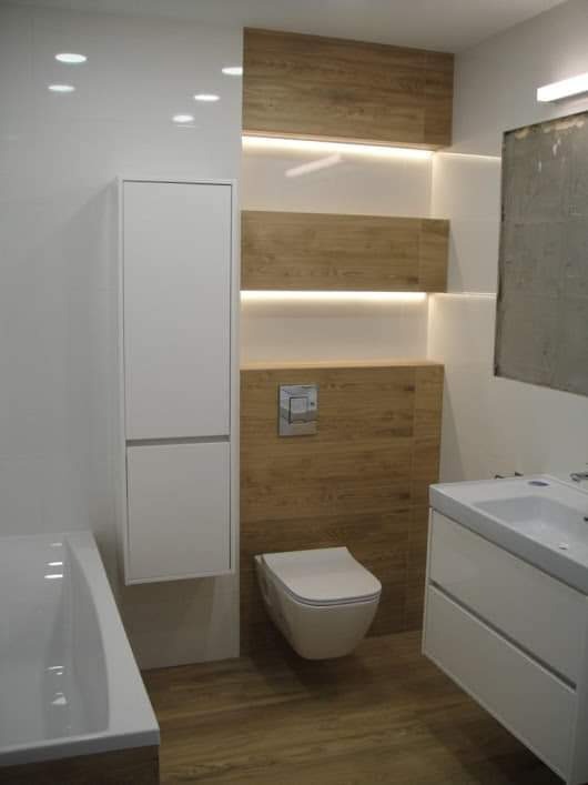 Nowoczesna łazienka z białymi meblami, drewnopodobnymi panelami ściennymi z oświetleniem LED, wiszącą toaletą i białą wanną.