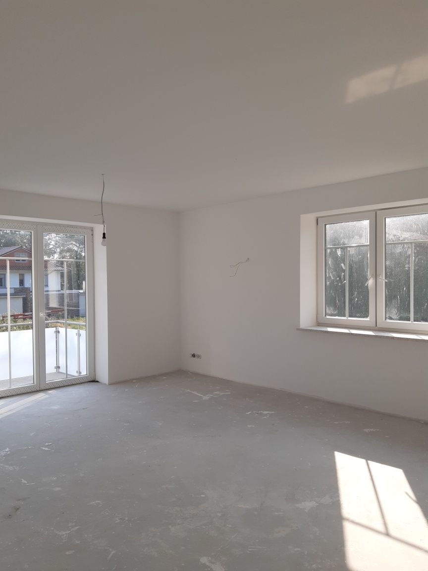 Pusty, jasny pokój w stanie deweloperskim z dużym oknem i wyjściem na balkon, surowy beton na podłodze, widoczne ślady po pracach wykończeniowych.