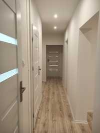 Wąski korytarz po remoncie z jasnymi ścianami, drewnopodobną podłogą i białymi drzwiami z poziomymi przeszkleniami, oświetlony dwoma punktowymi lampami sufitowymi.