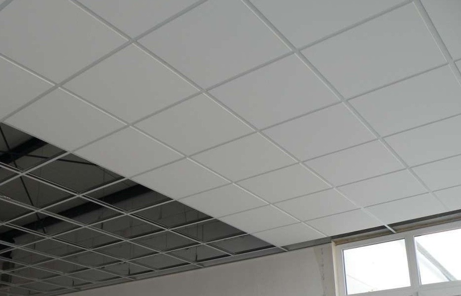 Częściowo zamontowany biały sufit podwieszany z widoczną metalową konstrukcją i oknem w tle.