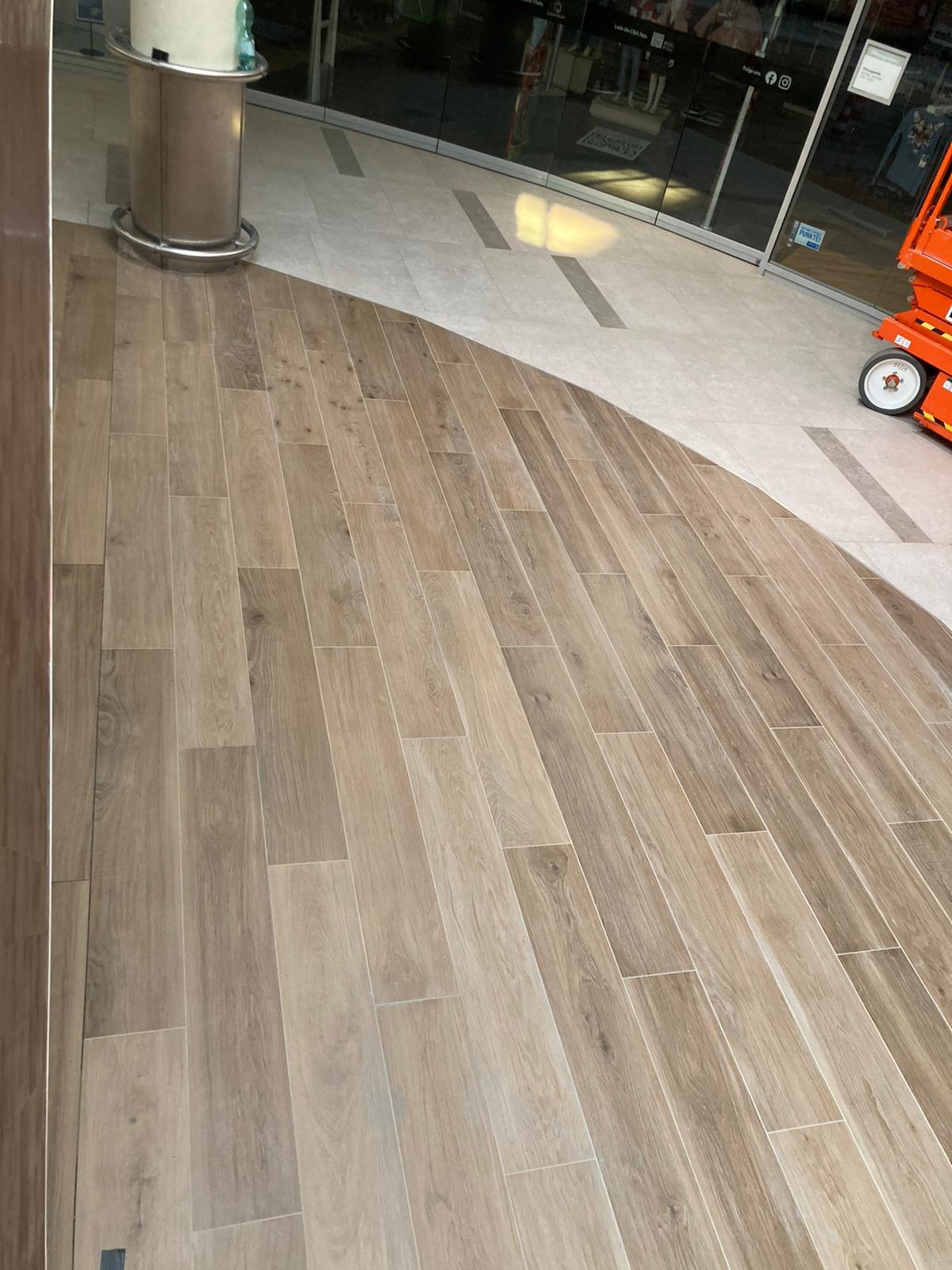 Ułożona pod kątem jasnobrązowa podłoga z płytek imitujących drewno przechodząca w szare płytki, widoczny fragment podnośnika koszowego przy szklanych drzwiach wejściowych.