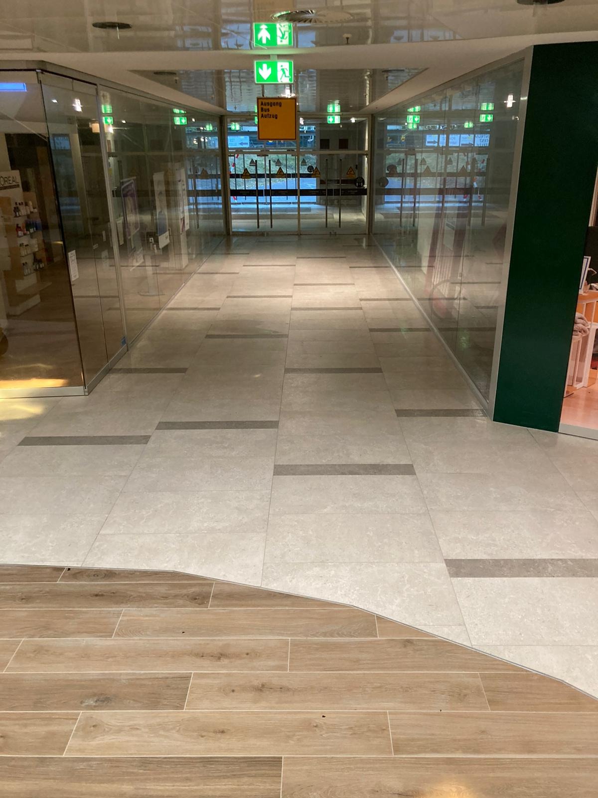 Połączenie płytek ceramicznych w odcieniach beżu z panelami podłogowymi imitującymi drewno w korytarzu komercyjnym, widoczne szklane ściany i oznaczenia ewakuacyjne.