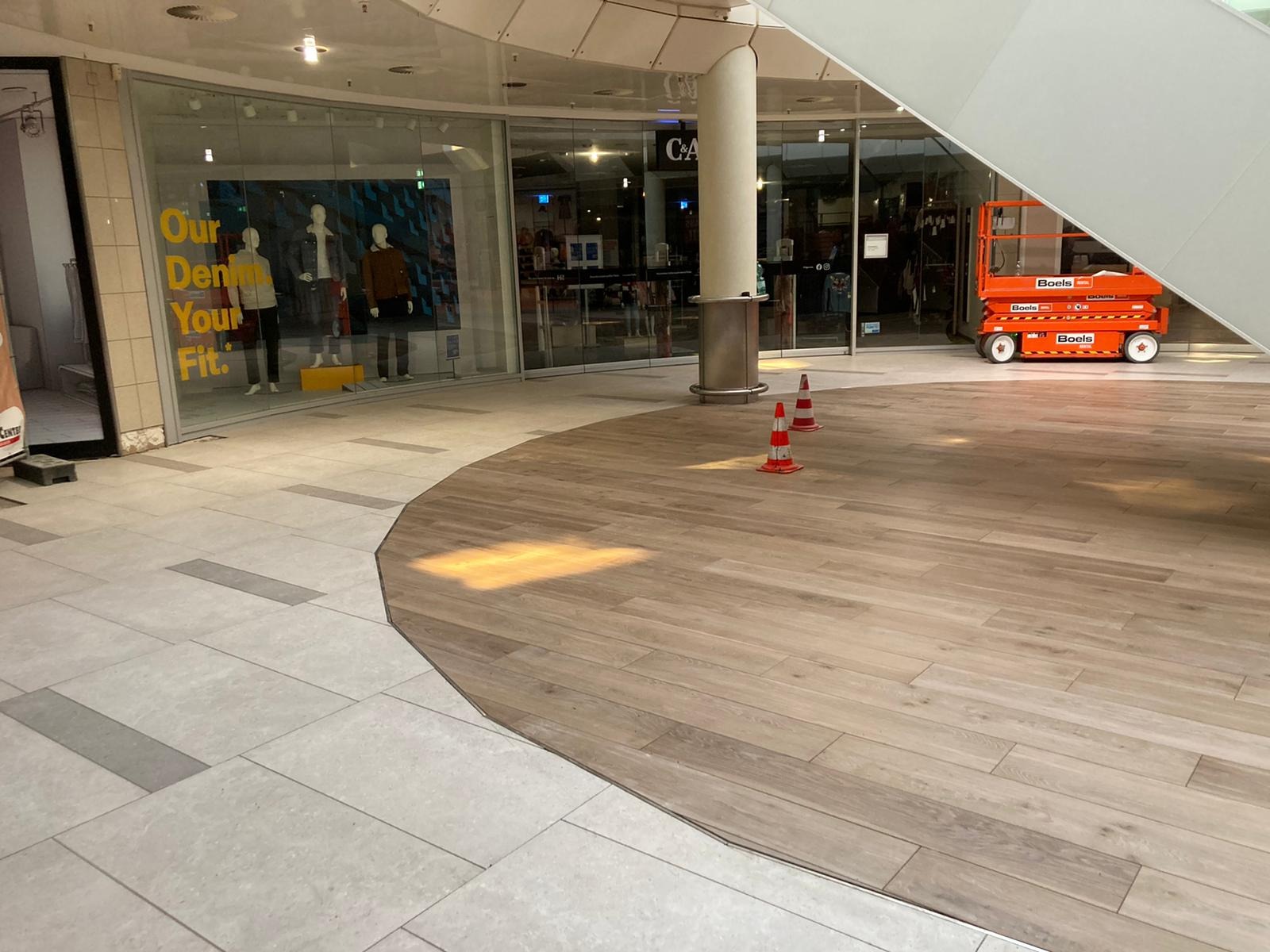 Nowa podłoga w galerii handlowej, połączenie płytek imitujących drewno i jasnych płytek, pomarańczowy podnośnik w tle, dwa pachołki ostrzegawcze.
