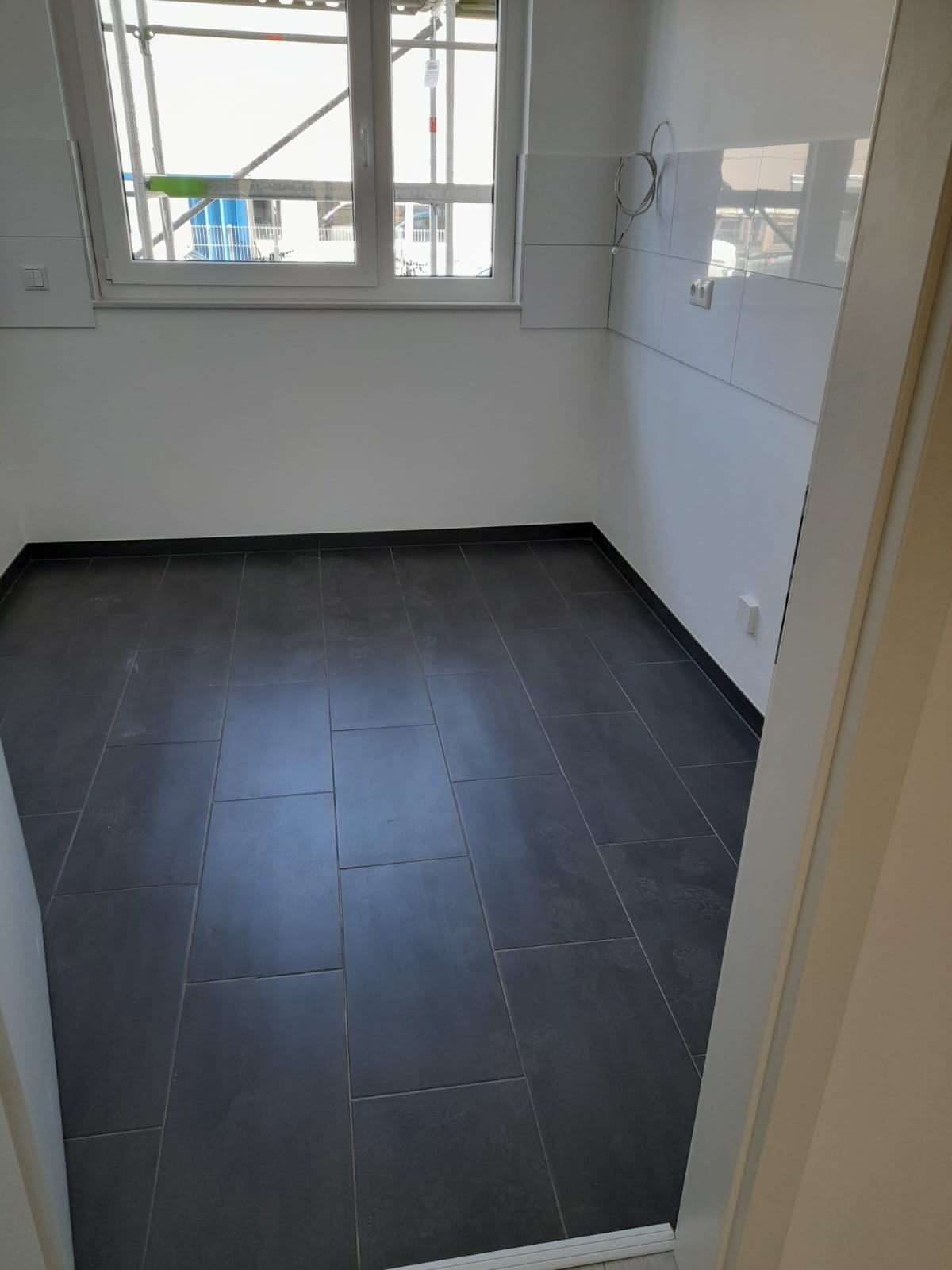 Ułożona, ciemnoszara podłoga z płytek w pustym pomieszczeniu z białymi ścianami i oknem, widok zza otwartych drzwi.