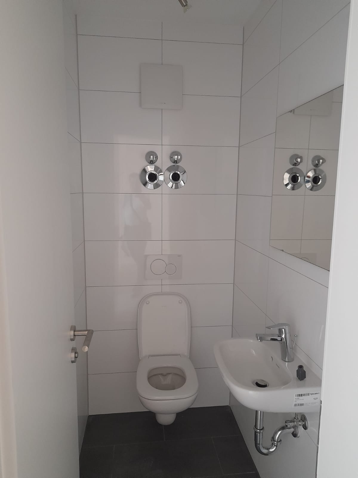 Nowoczesna, minimalistyczna toaleta z białymi płytkami, wiszącą umywalką i chromowaną armaturą, widoczny fragment lustra i wentylator na ścianie.